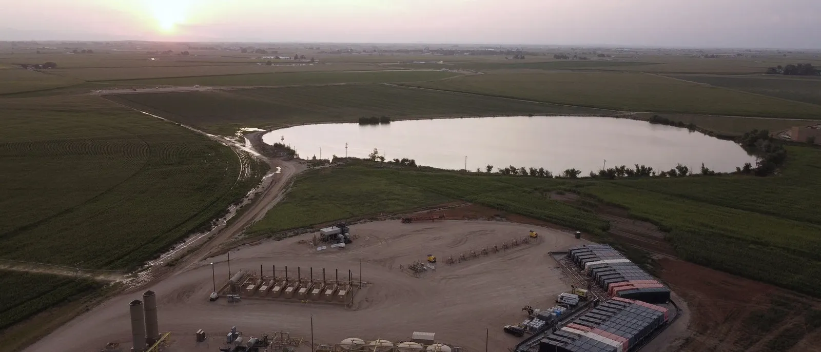 Aerial photograph at dusk of a PWR site in the DJ Basin: treatment skids, storage tanks and wellhead equipment beside a lined water impoundment, surrounded by irrigated farmland.